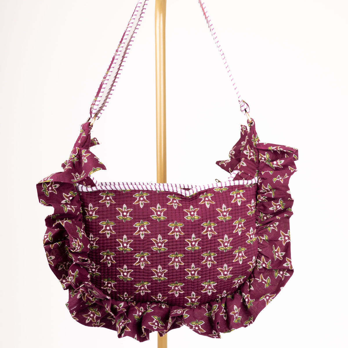 A ruffled shoulder bag in a deep purple fabric with a repeating gold fleur-de-lis pattern, hanging from a gold stand against a plain white background.