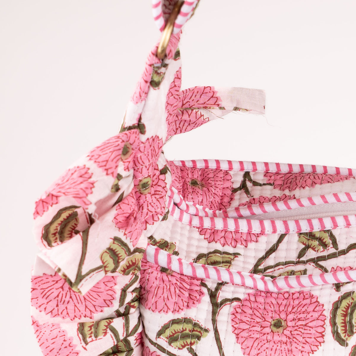 Close-up of a fabric handbag or pouch with a pink and green floral block-print pattern on a white background, featuring a pink and white striped zipper and strap.