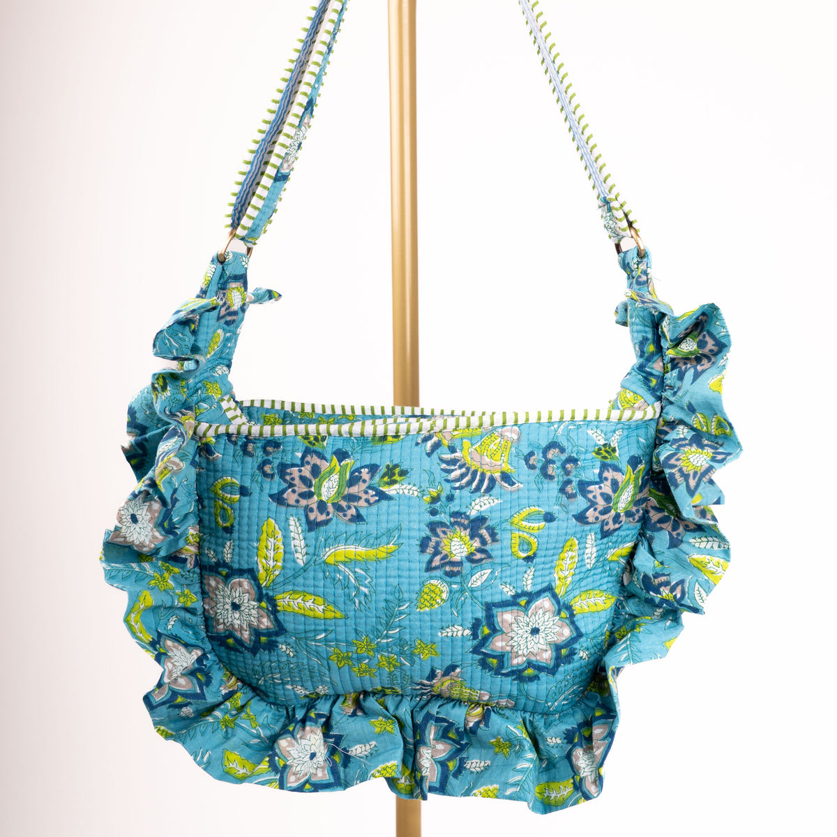 A ruffled shoulder bag with a blue, green, and white floral pattern, hanging from a gold-colored metal stand against a plain white background.