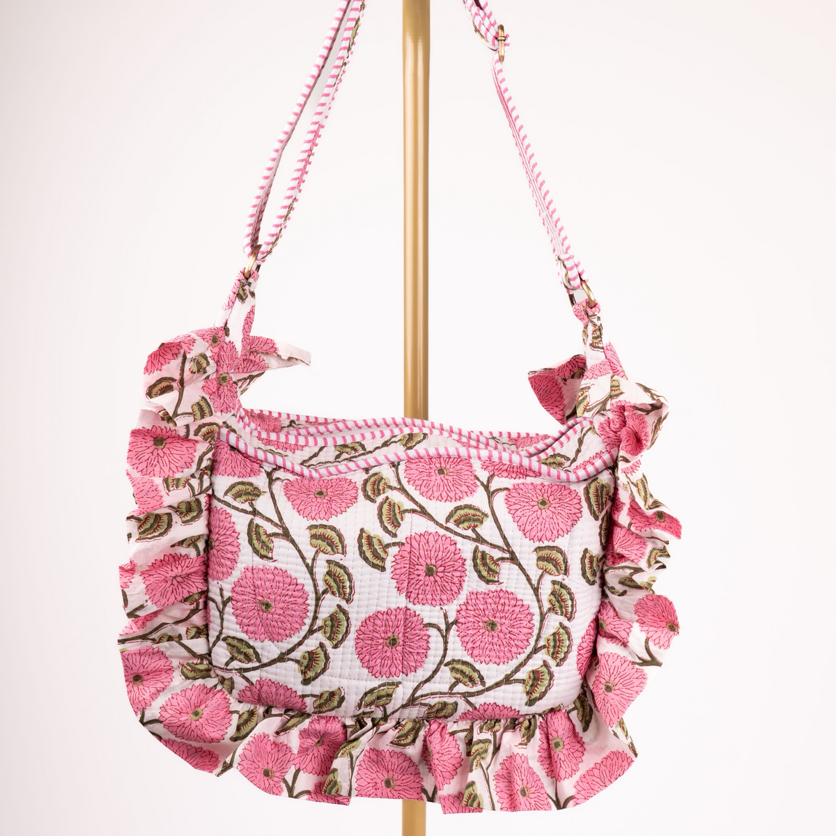 A pink floral shoulder bag with ruffled trim, hanging from a gold stand against a plain white background.