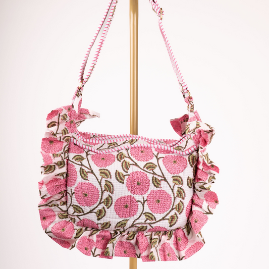 A pink floral shoulder bag with ruffled trim, hanging from a gold stand against a plain white background.