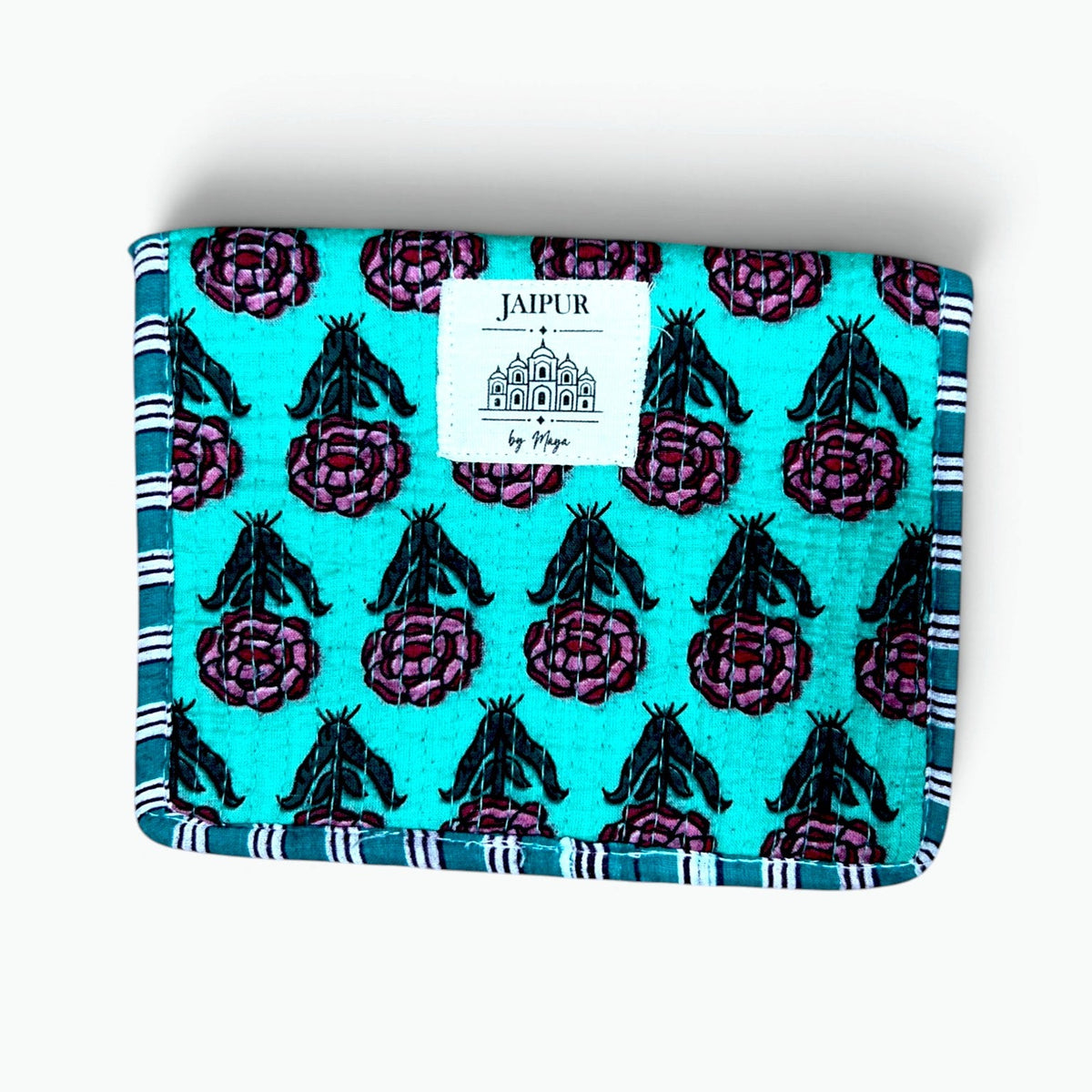 A square fabric pouch or notebook cover featuring a turquoise background with a repeating pattern of dark purple thistle flowers and black leaves, finished with a striped blue and white border. A white paper label on the front reads 'JOURNEE' with a small tree logo.